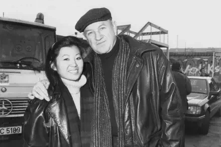Black and white image of Gene Hackman posing with a woman, both smiling. They stand in front of a vehicle, conveying a warm and friendly atmosphere. HD desktop wallpaper.