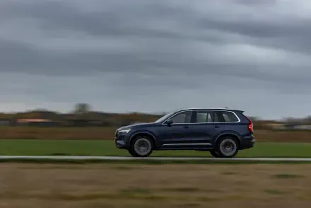 A sleek Volvo XC90 Facelift glides along a road against a dramatic sky, showcasing its modern design in this stunning 4K Ultra HD wallpaper.