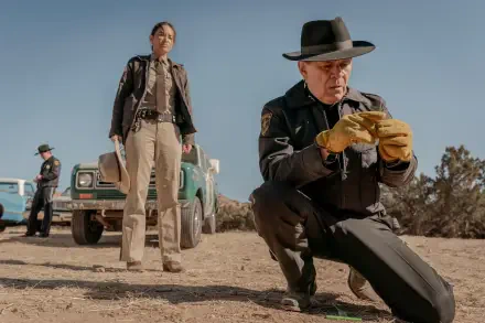 A scene from the TV show Dark Winds, featuring two law enforcement officers investigating a crime scene in a desert landscape, with a vintage vehicle in the background. 
