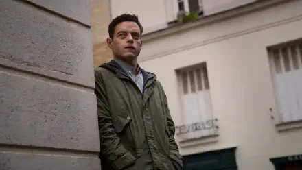 Rami Malek stands pensively against a wall in a scene from The Amateur (2025). This captivating image serves as an 8K Ultra HD desktop wallpaper or background.