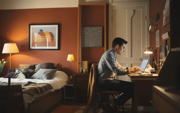 A digital nomad works focused at a desk in a cozy, warmly-lit room, with a bed and artwork in the background, showcasing a blend of productivity and comfort.