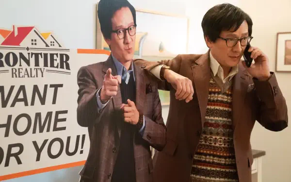 A scene from the movie Love Hurts, featuring a character in a brown suit standing next to a life-sized cardboard cutout, holding a phone and interacting with a Frontier Realty sign.