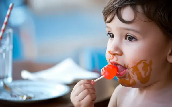 A young child enjoys a bright red lollipop, with playful expression and a smeared candy face, set against a softly blurred background, captured in vibrant 4K Ultra HD.