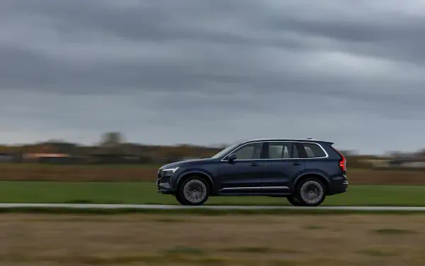 A sleek Volvo XC90 Facelift glides along a road against a dramatic sky, showcasing its modern design in this stunning 4K Ultra HD wallpaper.