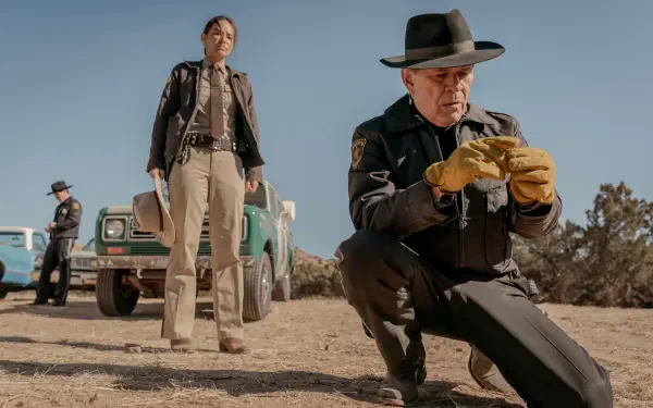A scene from the TV show Dark Winds, featuring two law enforcement officers investigating a crime scene in a desert landscape, with a vintage vehicle in the background. 