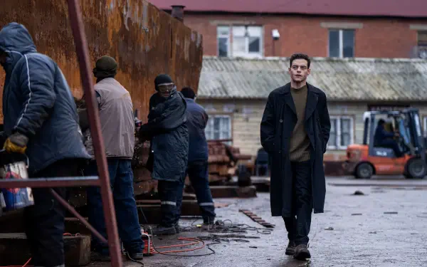 A scene from the movie The Amateur (2025) featuring Rami Malek, showcasing a gritty industrial setting with several workers and a lone figure walking amidst them. 8K Ultra HD quality.
