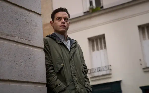 Rami Malek stands pensively against a wall in a scene from The Amateur (2025). This captivating image serves as an 8K Ultra HD desktop wallpaper or background.