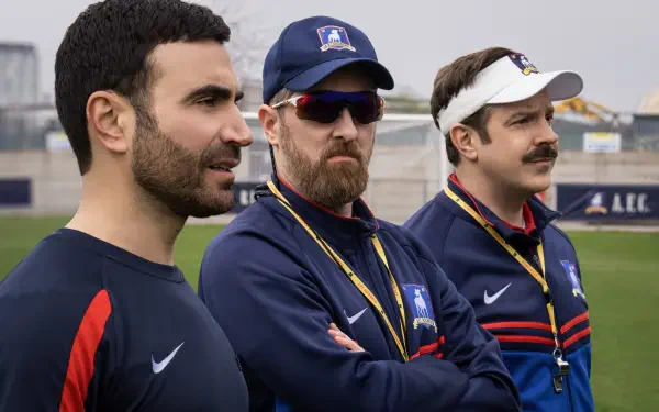 A scene from the TV show Ted Lasso, featuring Jason Sudeikis, Brendan Hunt, and Brett Goldstein as they observe a practice on the field, portrayed in vibrant 8K Ultra HD.