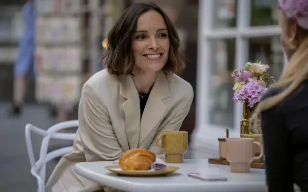 A scene from the TV show Ted Lasso featuring Jodi Balfour, smiling at a table with coffee and pastries, surrounded by flowers in an inviting café setting. 8K Ultra HD wallpaper.