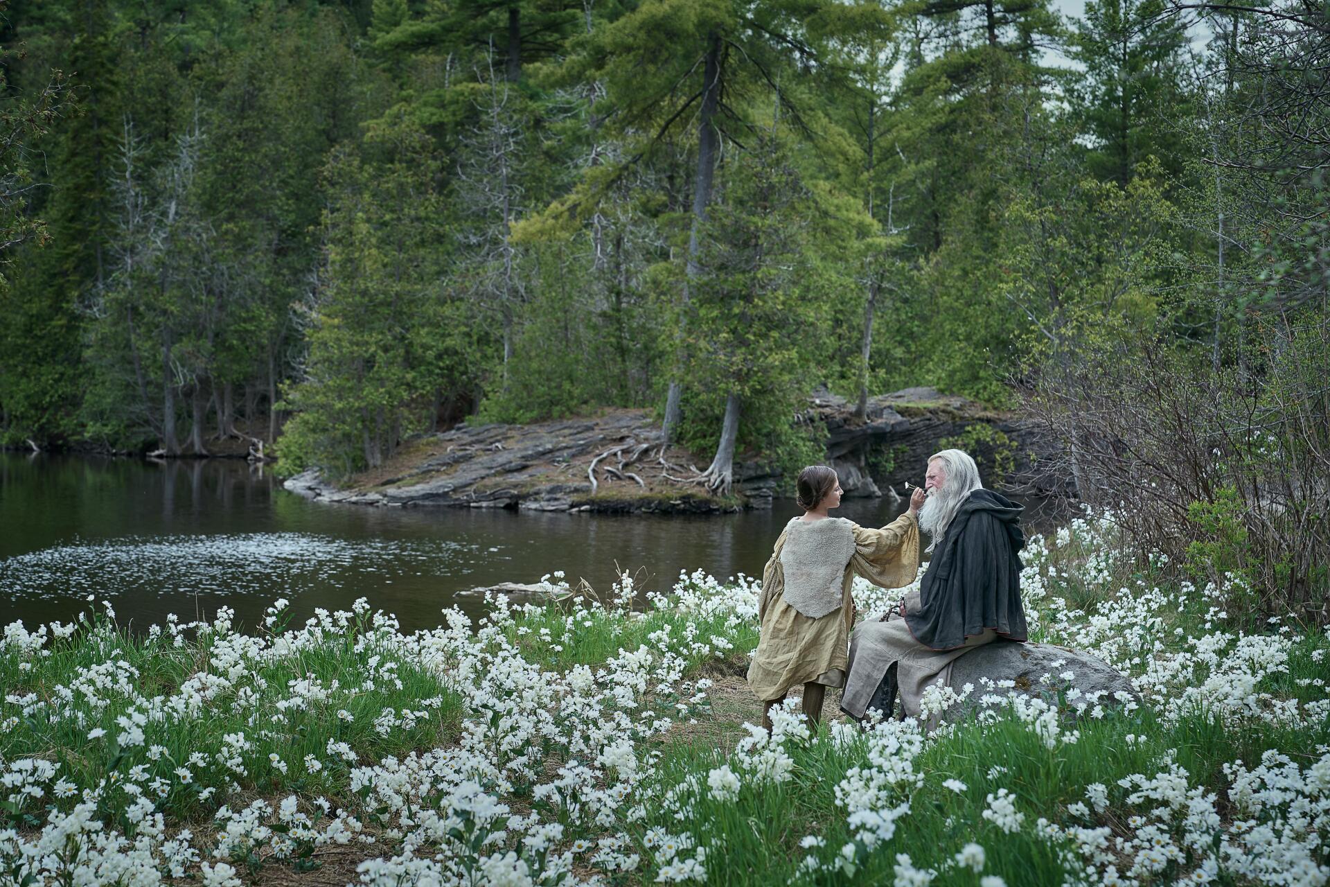 4K Ultra HD PC desktop wallpaper/background: scene from the movie Frankenstein (2025) — two figures share a quiet moment by a lake amid white wildflowers and dense evergreen woods.