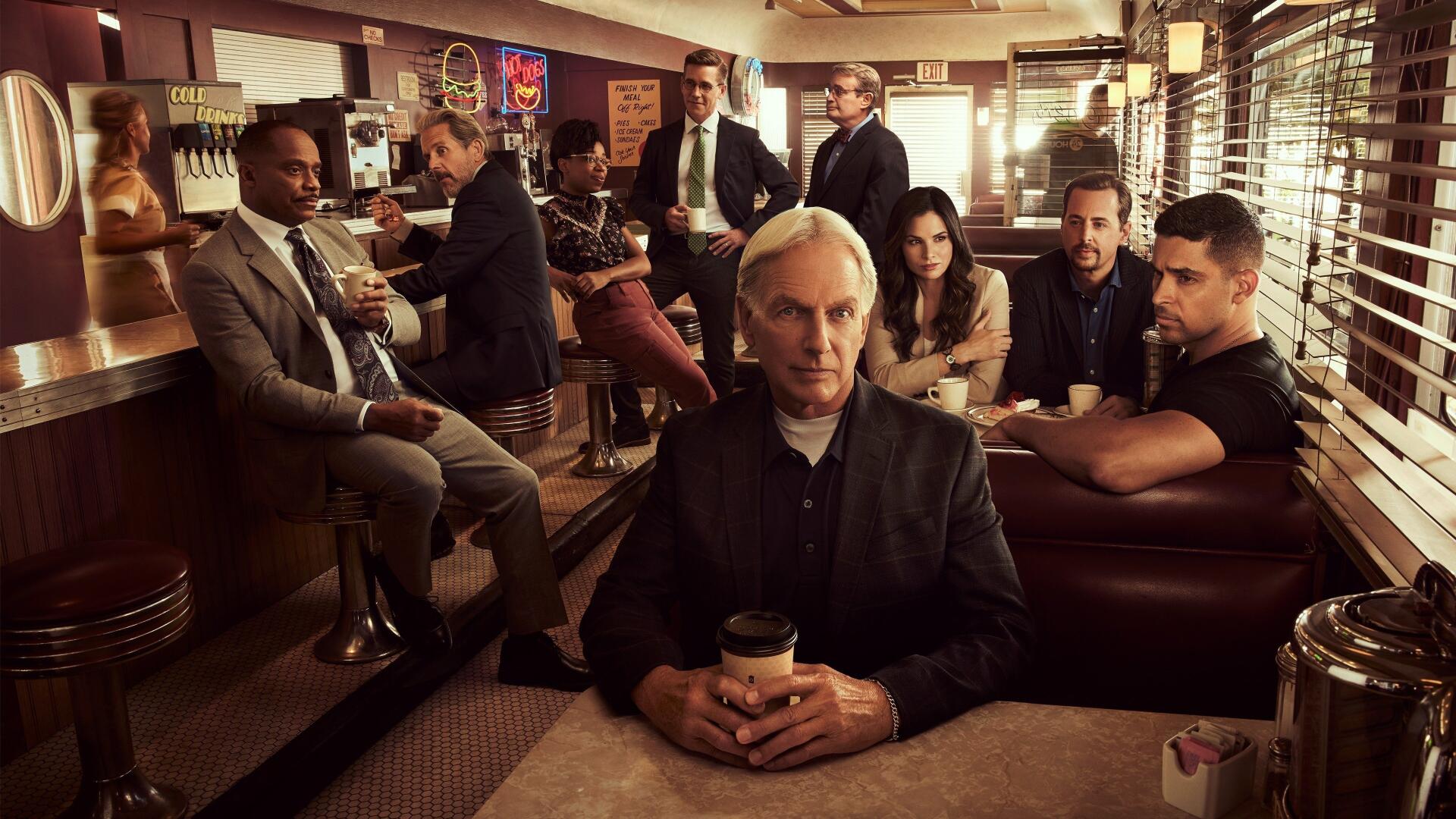 NCIS-themed 4K Ultra HD PC desktop wallpaper showing an ensemble in a dim diner; lead figure with coffee in foreground, team clustered behind under warm, cinematic lighting.
