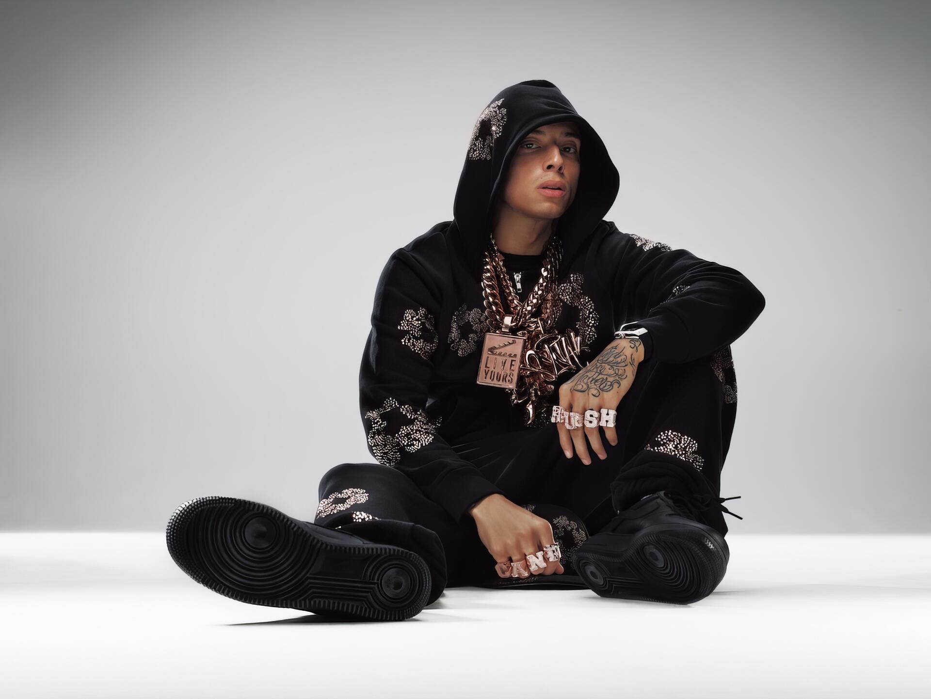 HD desktop wallpaper of a British rapper (identity unknown) in a black hoodie, heavy chains and rings, seated on a white studio floor, looking ahead.