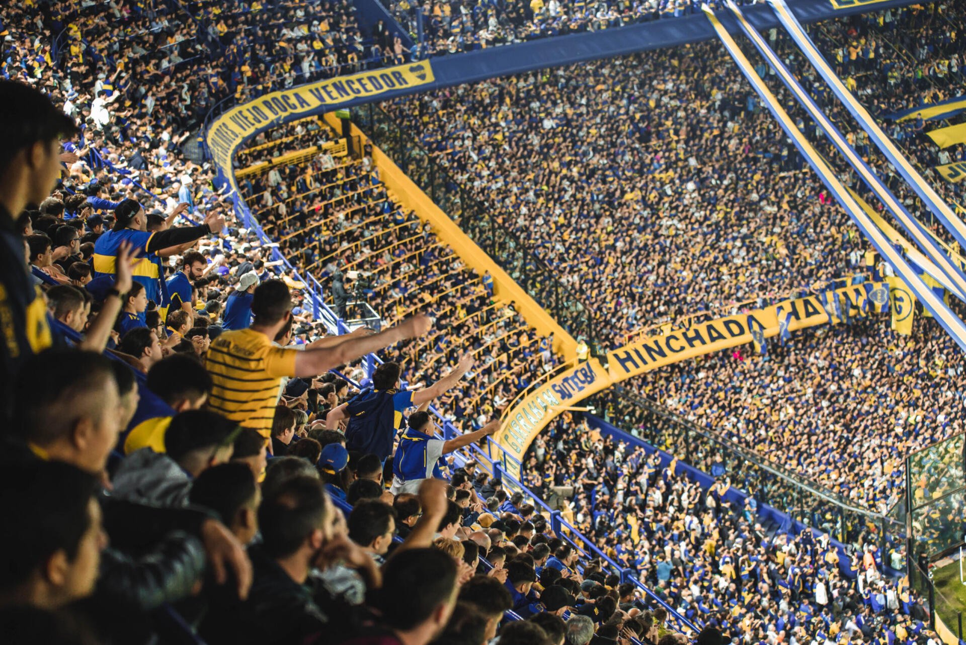 Panoramic 2K Quad HD desktop wallpaper of a packed Boca Juniors crowd in Argentina, fans in blue and yellow filling steep stands and waving banners.