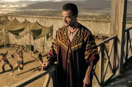 HD PC desktop wallpaper/background from the TV show Spartacus: House of Ashur — a robed man on a balcony overlooking a sunlit arena where gladiators spar beside tents and stone walls.