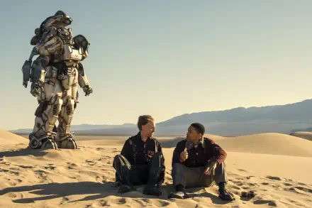 Fallout (TV Show) 2K Quad HD PC desktop wallpaper: two characters sit on desert dunes sharing food while a towering Power Armor suit stands behind, pale sky and distant mountains.