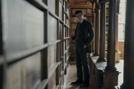 TV show scene: Young Sherlock detective stands in a shadowed library aisle, hands in coat, moody 2K Quad HD PC desktop wallpaper background.