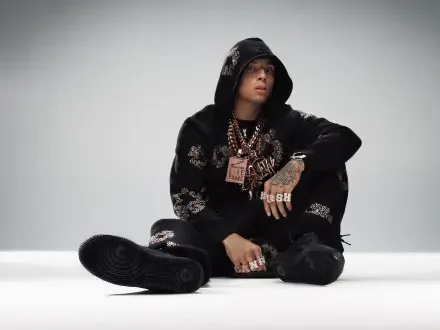 HD desktop wallpaper of a British rapper (identity unknown) in a black hoodie, heavy chains and rings, seated on a white studio floor, looking ahead.