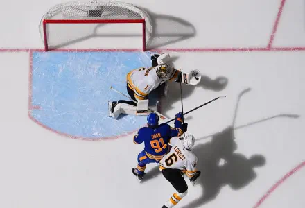 Overhead hockey photography: NHL action between Buffalo Sabres and Boston Bruins at the crease — goalie and two skaters with long shadows on the ice. 2K Quad HD PC desktop wallpaper background.