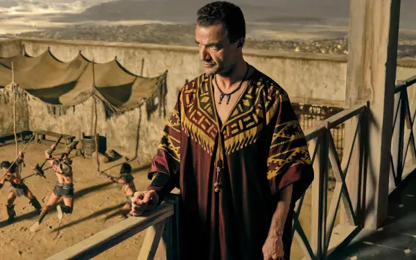 HD PC desktop wallpaper/background from the TV show Spartacus: House of Ashur — a robed man on a balcony overlooking a sunlit arena where gladiators spar beside tents and stone walls.