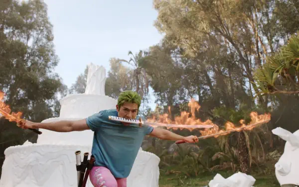 2K Quad HD PC desktop wallpaper: Roronoa Zoro from the TV show One Piece (Live Action) grips a sword in his mouth as flaming blades arc beside stone sculptures in a forest.