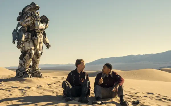 Fallout (TV Show) 2K Quad HD PC desktop wallpaper: two characters sit on desert dunes sharing food while a towering Power Armor suit stands behind, pale sky and distant mountains.