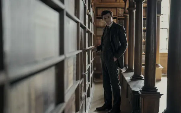TV show scene: Young Sherlock detective stands in a shadowed library aisle, hands in coat, moody 2K Quad HD PC desktop wallpaper background.