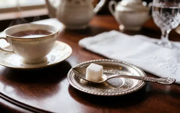 5K Ultra HD PC desktop wallpaper: a sugar cube on a spoon on a silver plate beside a teacup and linen napkin on a polished wood table.