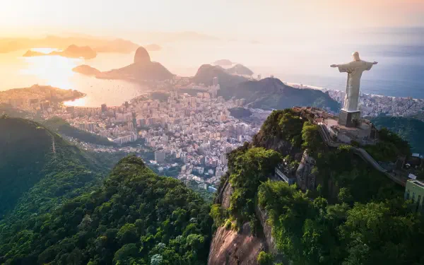 4K aerial landscape of Rio de Janeiro at sunset, Christ the Redeemer atop Corcovado overlooking the city, lush green hills and the coastline with Sugarloaf Mountain.