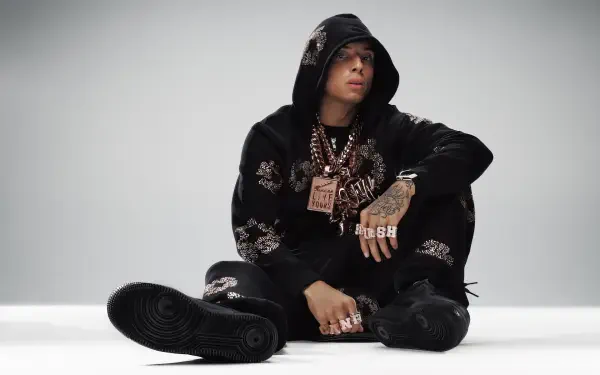 HD desktop wallpaper of a British rapper (identity unknown) in a black hoodie, heavy chains and rings, seated on a white studio floor, looking ahead.