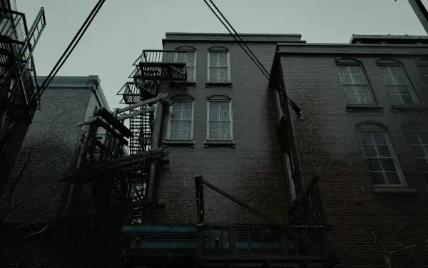 Resident Evil Requiem 2K Quad HD PC desktop wallpaper: moody desaturated alley of brick buildings with fire escapes, overhead wires and a gloomy sky.