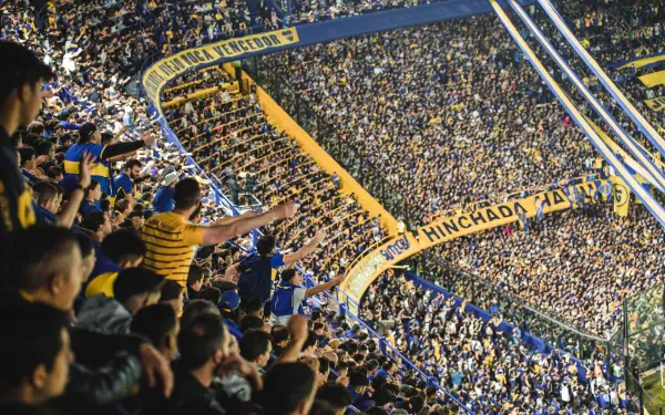Panoramic 2K Quad HD desktop wallpaper of a packed Boca Juniors crowd in Argentina, fans in blue and yellow filling steep stands and waving banners.