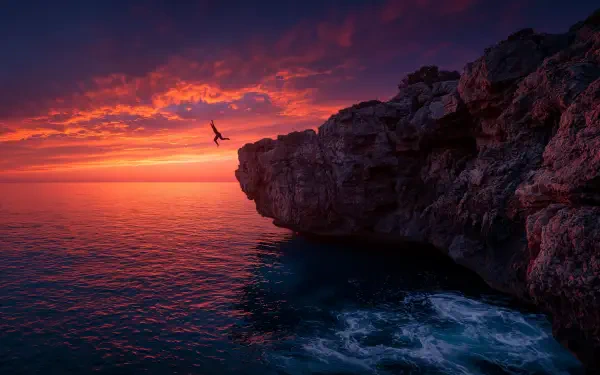 Silhouette jumping from a rocky cliff into the ocean at a summer sunset, vibrant orange sky and waves — 2K Quad HD PC desktop wallpaper and background.