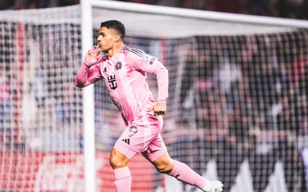 HD desktop wallpaper of an Inter Miami CF soccer forward in pink kit celebrating a goal by the net, sprinting with a finger to his lips amid a packed stadium.