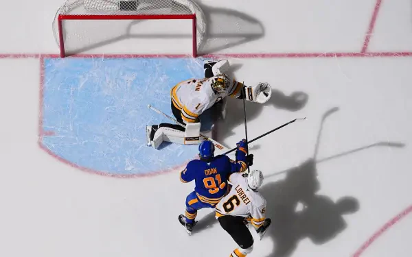 Overhead hockey photography: NHL action between Buffalo Sabres and Boston Bruins at the crease — goalie and two skaters with long shadows on the ice. 2K Quad HD PC desktop wallpaper background.