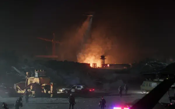Resident Evil Requiem 2K Quad HD PC Desktop Wallpaper and Background — nighttime industrial ruin with massive fire, smoky sky, military vehicles and armed silhouettes in the foreground.