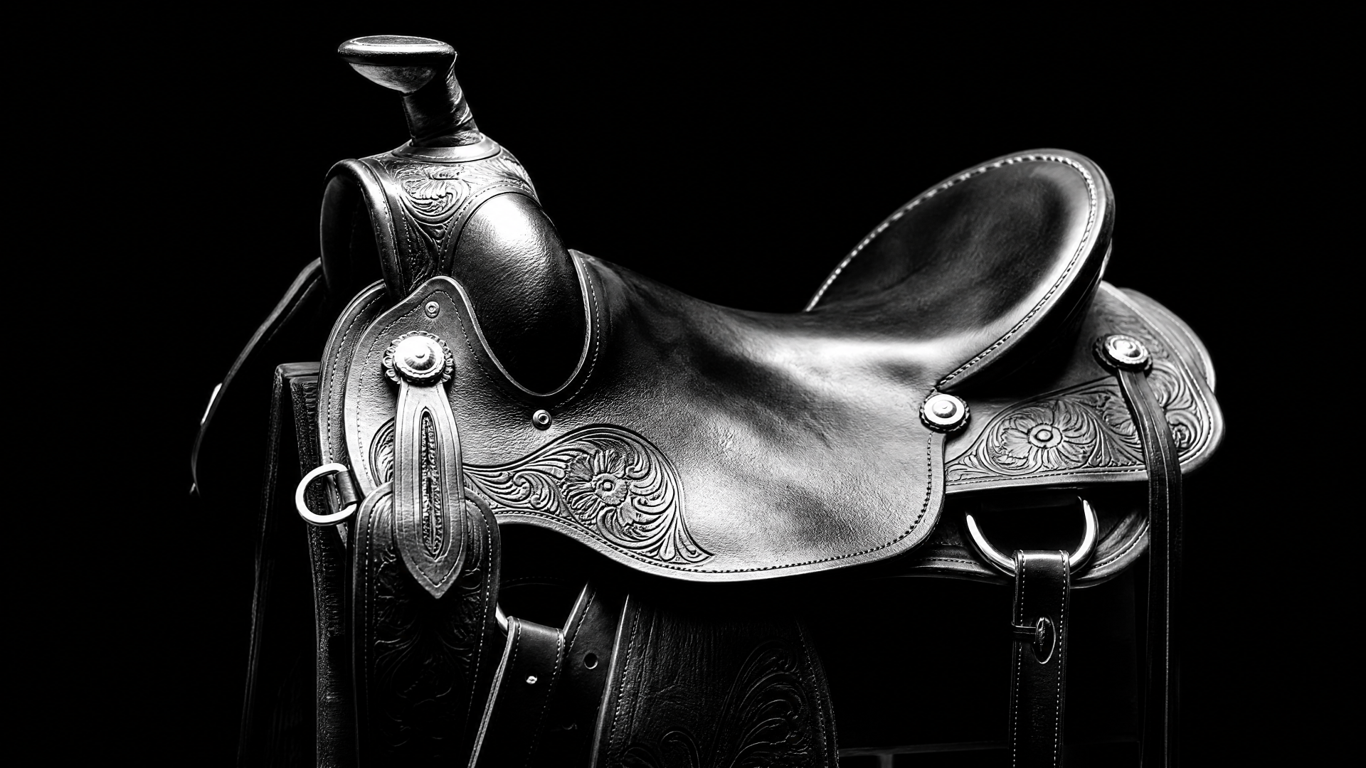 Ornately tooled leather Western saddle on a black background; high-contrast black-and-white 4K Ultra HD PC desktop wallpaper and background.