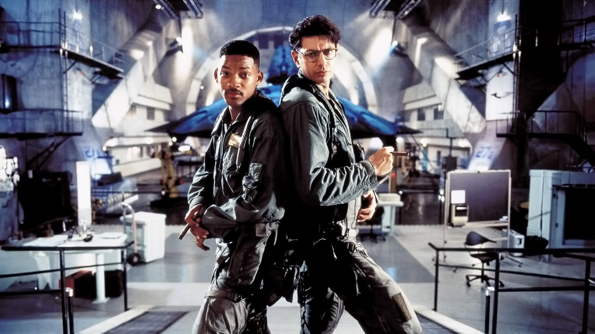 Independence Day 4K Ultra HD PC desktop wallpaper: two pilots in flight suits stand back-to-back in a giant military hangar, dramatic lighting and a glowing spacecraft in the background.