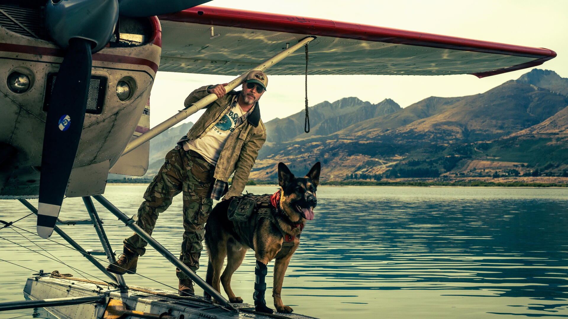 Pilot in flight jacket and sunglasses stands with a German Shepherd on a floatplane dock over a mountain-reflected lake, Heart of the Beast, 4K Ultra HD PC desktop wallpaper and background.