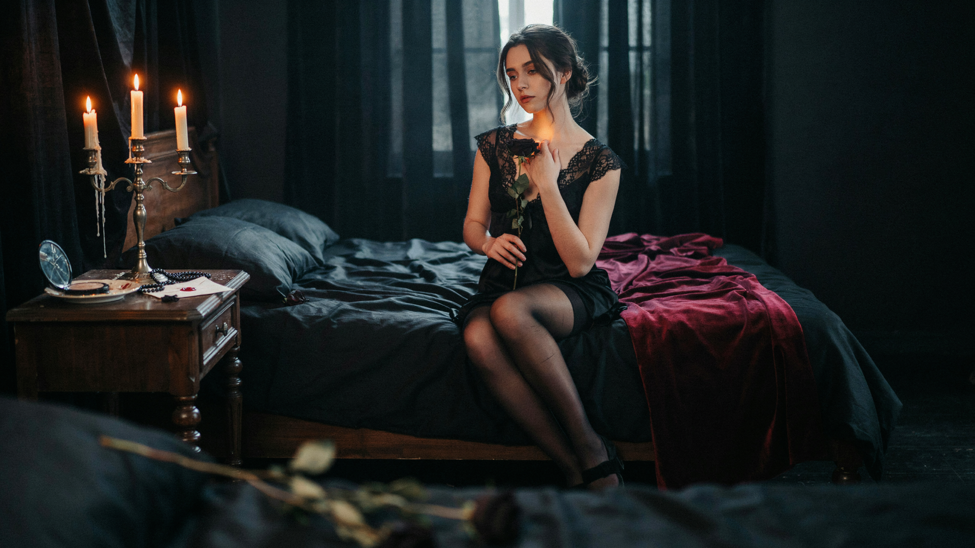 Dark coquette woman in a black dress sits on a bed by candlelight in a moody room — 2K Quad HD PC desktop wallpaper/background.