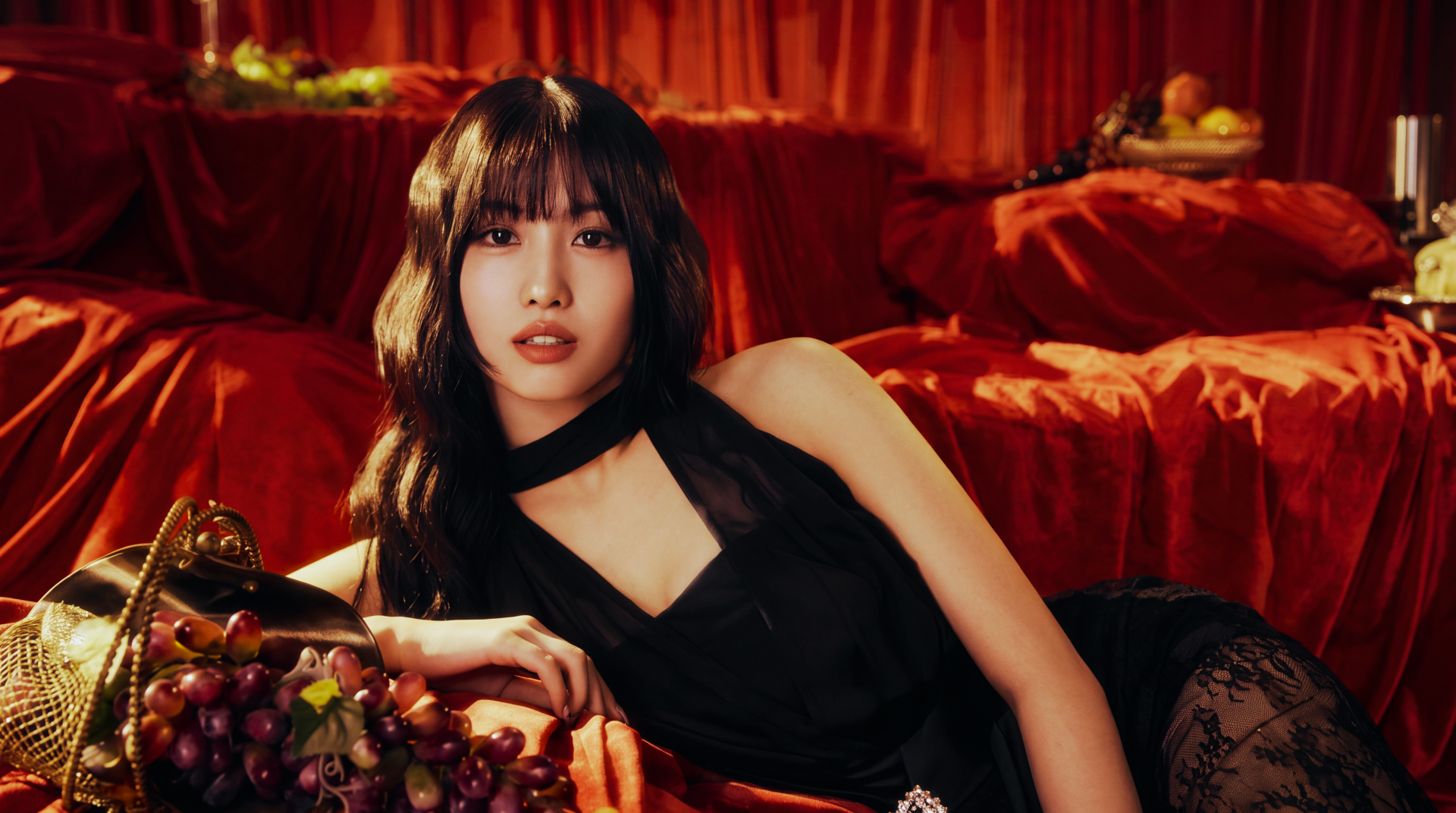5K desktop wallpaper of a Japanese model with a k-pop idol–inspired look reclining on red velvet in a black dress, grapes arranged in the foreground.