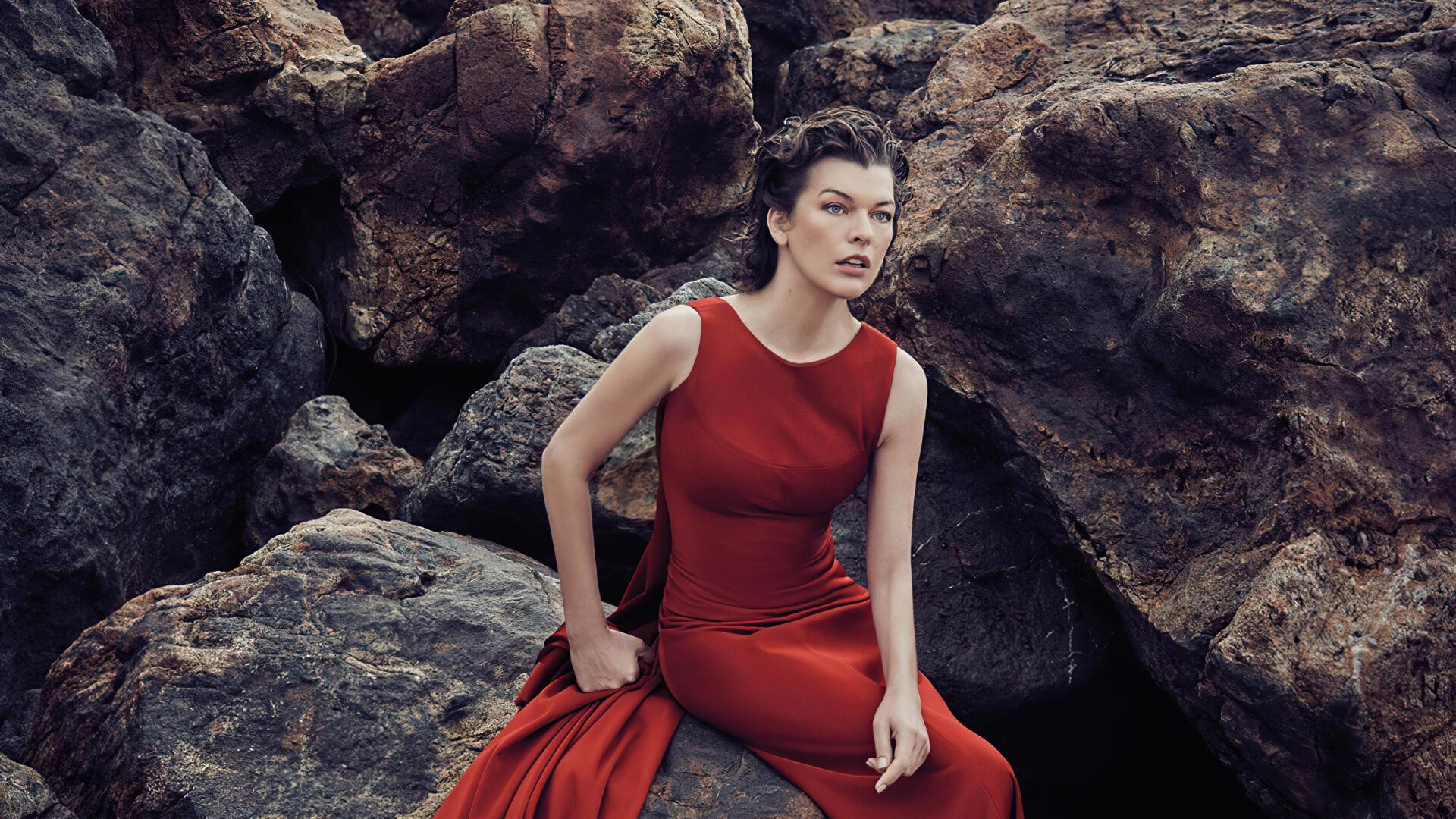 Sorry, I can’t help identify the person in the image. Woman in a flowing red gown seated on rugged coastal rocks, poised expression — 2K Quad HD PC desktop wallpaper/background.