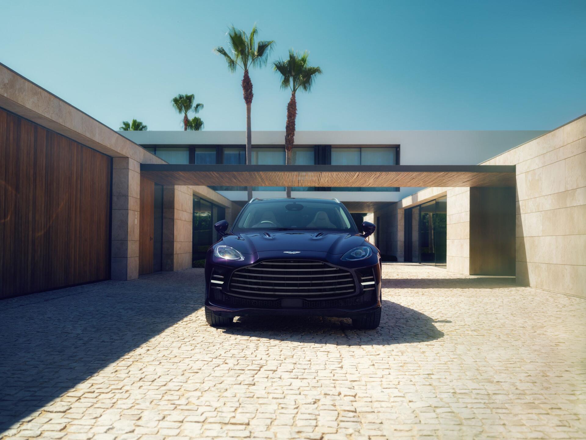 Front view of a blue Aston Martin DBX GT SUV in a sunlit modern courtyard — a luxury car presented as a 2K Quad HD PC desktop wallpaper.