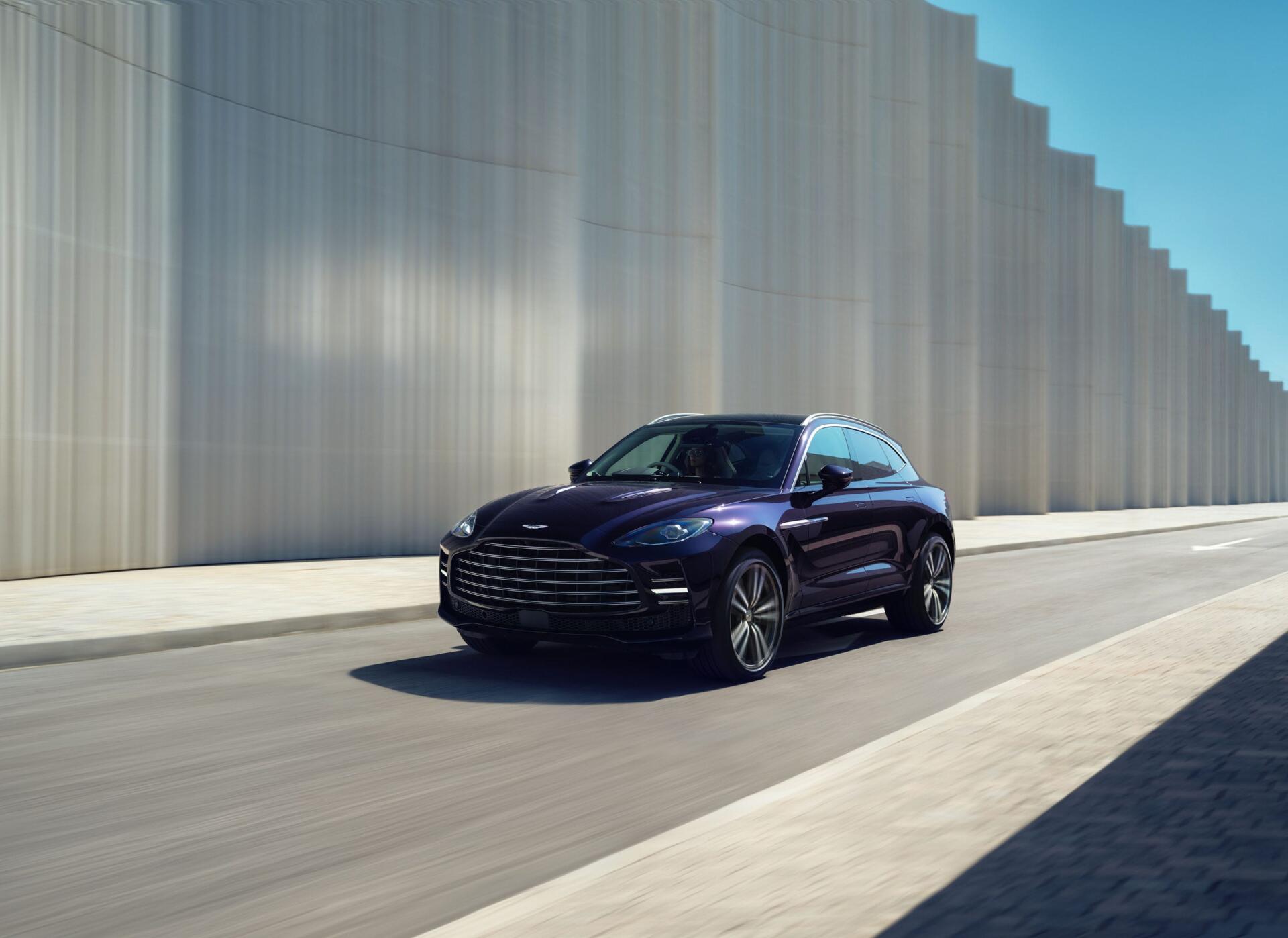Aston Martin DBX GT sedan driving past modern concrete walls, captured as a 2K Quad HD PC desktop wallpaper and background.