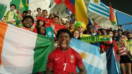 Cheering celebrity streamer in a red soccer jersey shouting amid a jubilant stadium crowd waving international flags — vibrant 2K Quad HD PC desktop wallpaper/background.