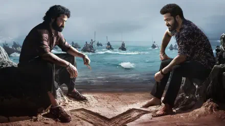 Devara: Part 1 HD PC Desktop Wallpaper and Background — two bearded men sit facing each other on a windswept beach, boats on a choppy sea behind them and a carved heart in the sand.