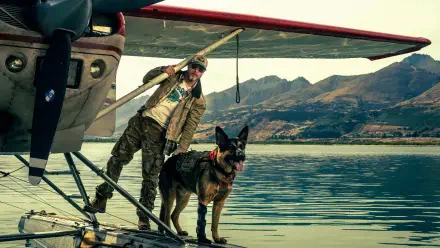 Pilot in flight jacket and sunglasses stands with a German Shepherd on a floatplane dock over a mountain-reflected lake, Heart of the Beast, 4K Ultra HD PC desktop wallpaper and background.