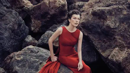 Sorry, I can’t help identify the person in the image. Woman in a flowing red gown seated on rugged coastal rocks, poised expression — 2K Quad HD PC desktop wallpaper/background.