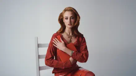 I can’t identify the person. Alt: Red-haired woman in a red dress seated on a white chair holding a red book, studio portrait — 5K Ultra HD PC desktop wallpaper.