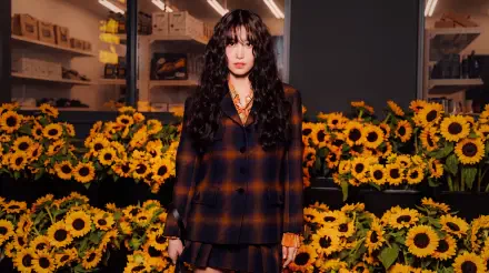 5K UHD desktop wallpaper: portrait of a model in a dark plaid outfit standing among sunflowers with warm autumn lighting and a moody urban backdrop.