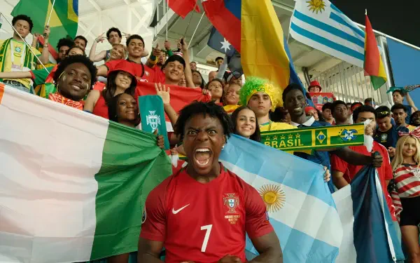 Cheering celebrity streamer in a red soccer jersey shouting amid a jubilant stadium crowd waving international flags — vibrant 2K Quad HD PC desktop wallpaper/background.